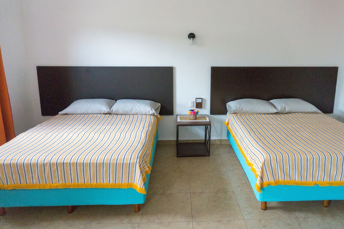 A hotel room with two beds, each with a striped bedspread and pillows, a small table with a clock and a vase of flowers, and a wall-mounted light fixture.