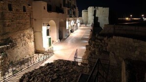 Vista nocturna de un edificio histórico de piedra con puertas en arco y un camino empedrado que conduce a una estructura fortificada al fondo.