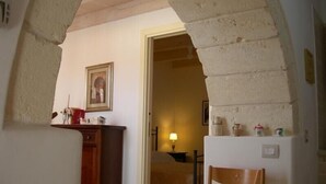 Una habitación con un escritorio de madera, una silla, una cama con cubrecama estampado y un acceso a otra habitación.