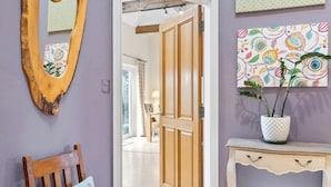Una stanza con una sedia e una porta in legno, un tavolino consolle bianco e una parete con uno specchio decorativo e opere d'arte a motivi.