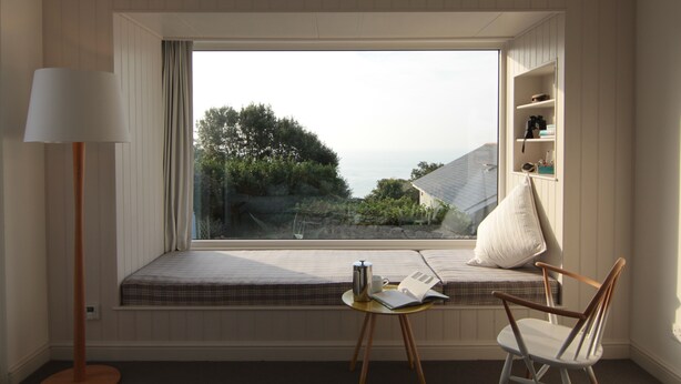 Ein Zimmer mit großem Fenster, Nachttisch mit Lampe, Stuhl und Regal mit Gegenständen.