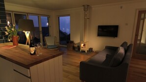 Ein gemütliches Wohnzimmer mit einer Couch, einem Holztisch mit Flasche und Gläsern, einem Kamin und einem Fernseher an der Wand.