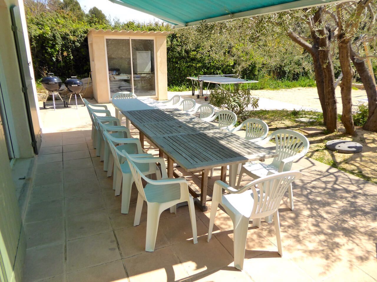 Een buitenterras met een lange tafel en witte stoelen, een barbecue en een overdekte zithoek.