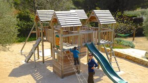 A wooden play structure with a slide and climbing net, surrounded by sand and greenery.