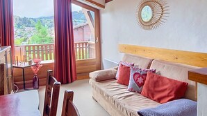 Een kamer met een houten hoofdeinde, een slaapbank, een eettafel met stoelen, een vaas met bloemen en uitzicht op een dorp door grote ramen.