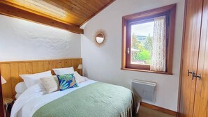 Een slaapkamer met een houten hoofdeinde, een bed met witte en kleurrijke kussens, een raam met uitzicht op groen, een radiator en een houten deur.