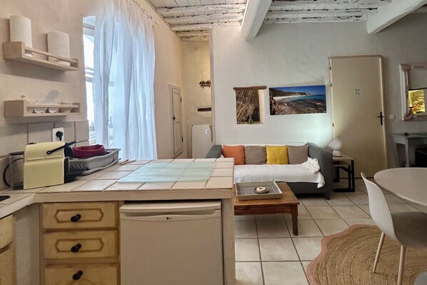 A compact living space with a kitchenette, a sofa with cushions, a dining table, and a view of the outdoors through a window.
