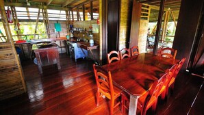 Un espace repas en bois avec une grande table et des chaises, un coin cuisine avec un comptoir et des appareils électroménagers, et une vue sur la verdure à l’extérieur.