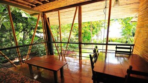 Un espace extérieur aménagé, abrité et doté de tables et de bancs en bois, entouré de bambous et de rampes en verre, offrant une vue sur un jardin luxuriant.