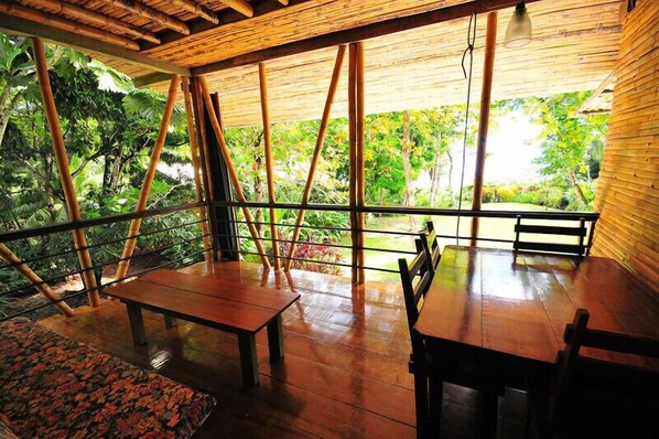 A covered outdoor seating area with wooden tables and benches, surrounded by bamboo poles and glass railings, overlooking a lush garden.
