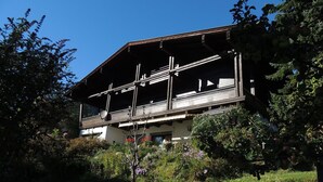 Une maison traditionnelle en bois, dotée d’un balcon, entourée d’une végétation luxuriante et d’arbres.