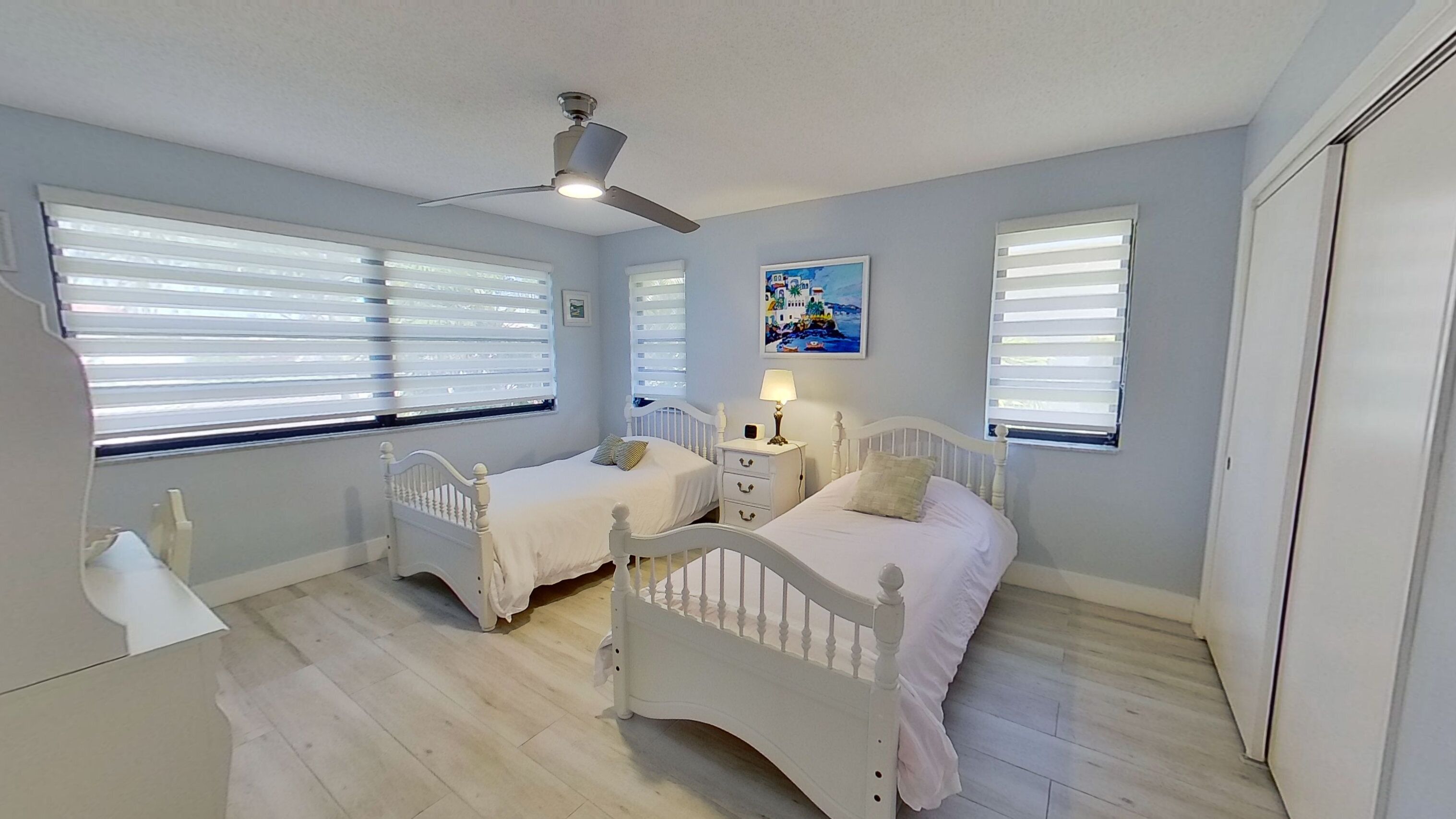 A bedroom with two white beds, a ceiling fan, and large windows with blinds.