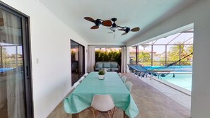 Un comedor con una mesa larga, sillas y un ventilador de techo. Se ve una piscina a través de amplios ventanales.