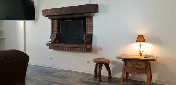 Een woonkamer met een haard van baksteen, een houten tafel met een lamp, een klein krukje en een aan de muur bevestigde televisie.