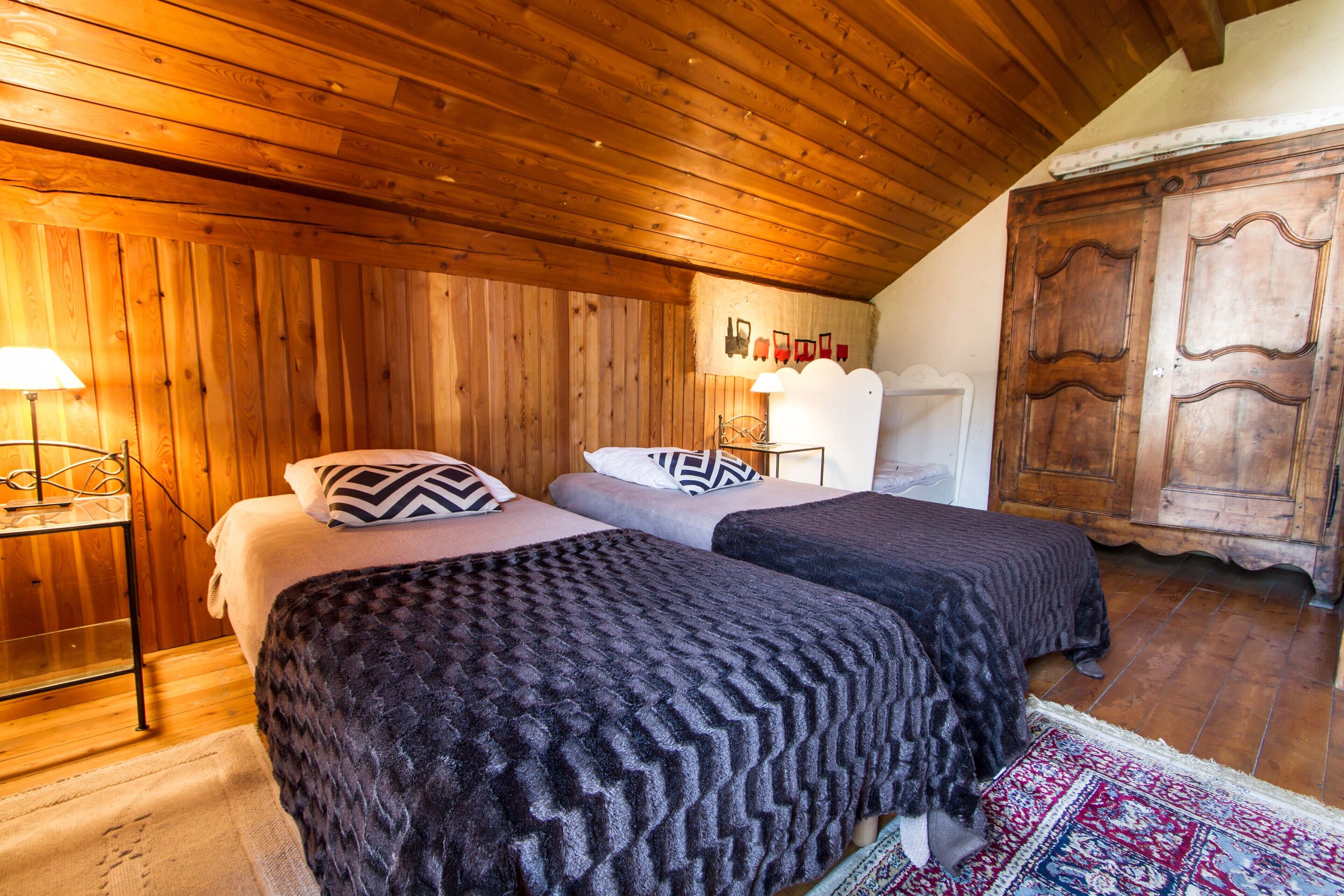 A room with two beds, wooden walls, a wooden ceiling, a wardrobe, and a patterned rug.