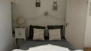 Een slaapkamer met een wit, versierd bed, een nachtkastje met een lamp en een deur.