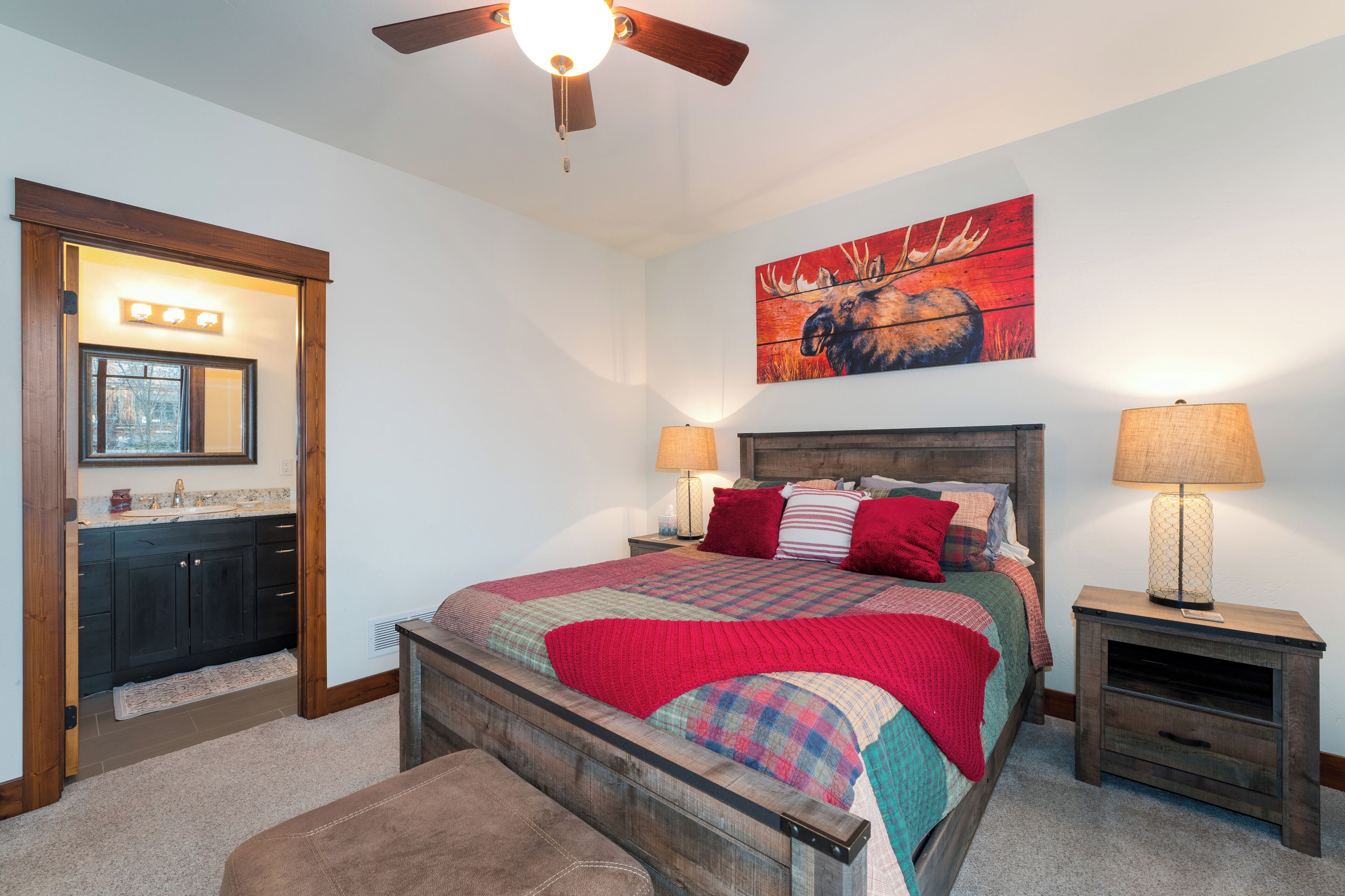 A bedroom with a wooden bed, a nightstand with a lamp, a painting of a moose, and a ceiling fan.