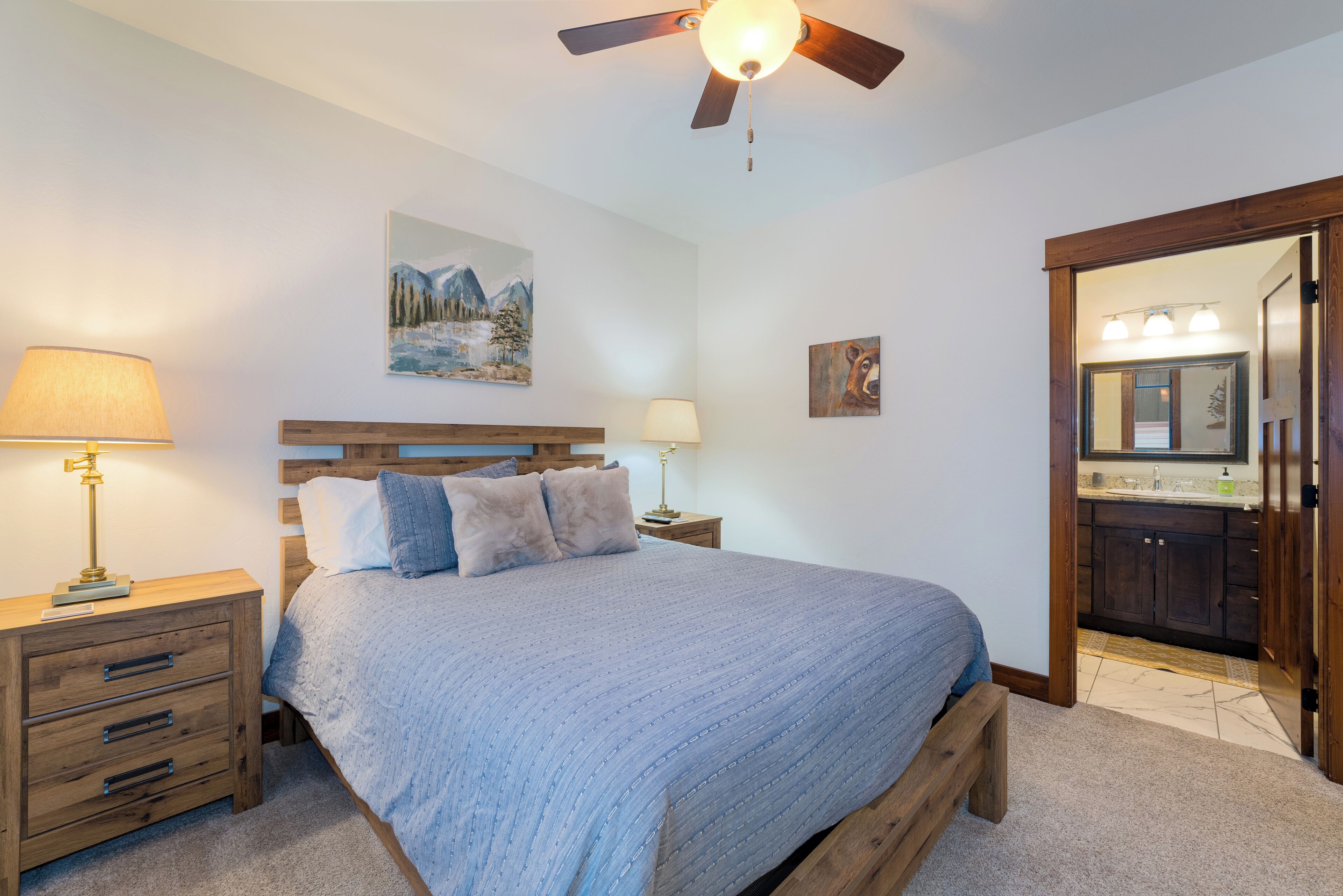 Un dormitorio con cama, cabecera de madera, mesitas de noche con lámparas, ventilador de techo, un cuadro y un baño visible a través de una puerta abierta.