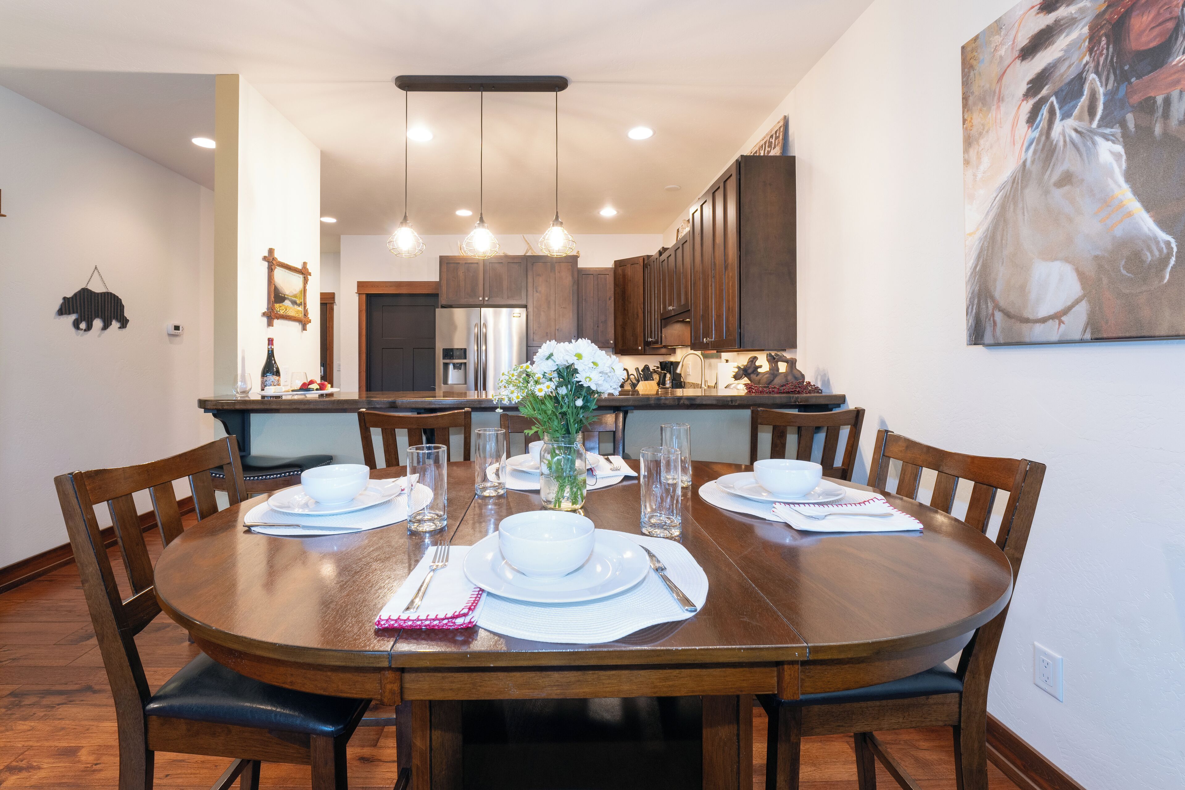 A dining area with a wooden table set for four, surrounded by chairs. There is a kitchen in the background with wooden cabinets and a refrigerator.