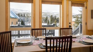 Un coin repas avec une table dressée pour quatre personnes, de grandes fenêtres équipées de stores et une vue sur des arbres et des montagnes couverts de neige.