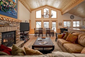 Ein gemütliches Wohnzimmer mit Kamin, Fernseher, Essbereich mit Tisch und Stühlen sowie einem großen Fenster mit Blick ins Freie.