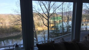 A room with a large window overlooking a river and trees. There is a bench with cushions and a table with a lamp and remote control.