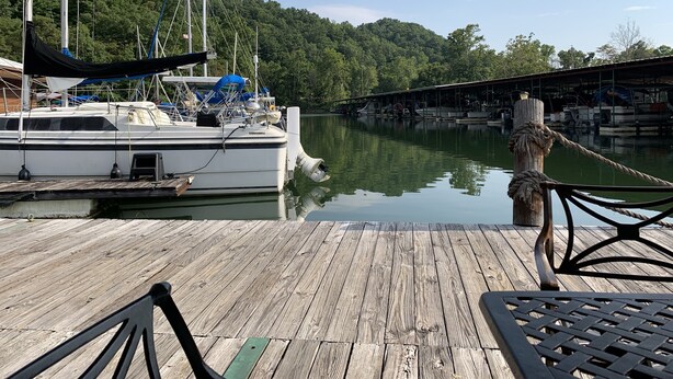 A wooden dock with a boat moored at the end, a covered seating area, and a forested hillside in the background.