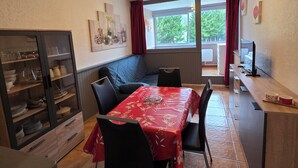 Un comedor con una mesa cubierta con mantel rojo, sillas negras, un armario de madera con puertas de cristal y vistas al exterior a través de una ventana con cortinas rojas.