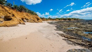Ein Strand mit blauem Himmel, felsiger Küste und spärlicher Vegetation.