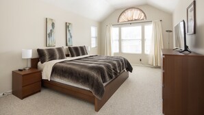 A bedroom with a bed, nightstand, lamp, ceiling fan, window with curtains, and artwork on the wall.