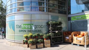 Edifici amb façana de vidre corbat i un cartell que diu "thefreshgrocer OBERT 24 HORES".