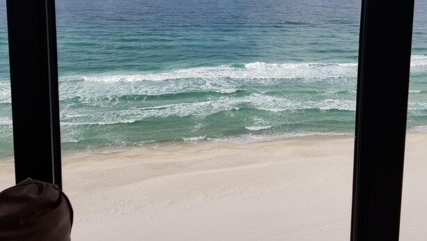 Une vue sur l’océan à travers de grandes fenêtres, avec une plage de sable visible au premier plan.
