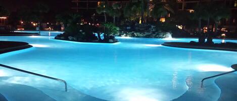 Una piscina de borde curvo, rodeada de palmeras y un edificio al fondo.