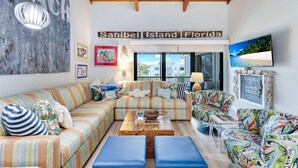 A cozy living room with striped sofas, a wooden coffee table, and a large window offering a view of the outside. - Coquina Beach 3G- Gorgeous 2bd, plus loft, Sanibel beach condo! (Sanibel)