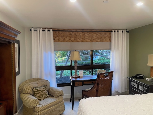 A room with a bed, a desk with a lamp, a chair, and a view of greenery outside.