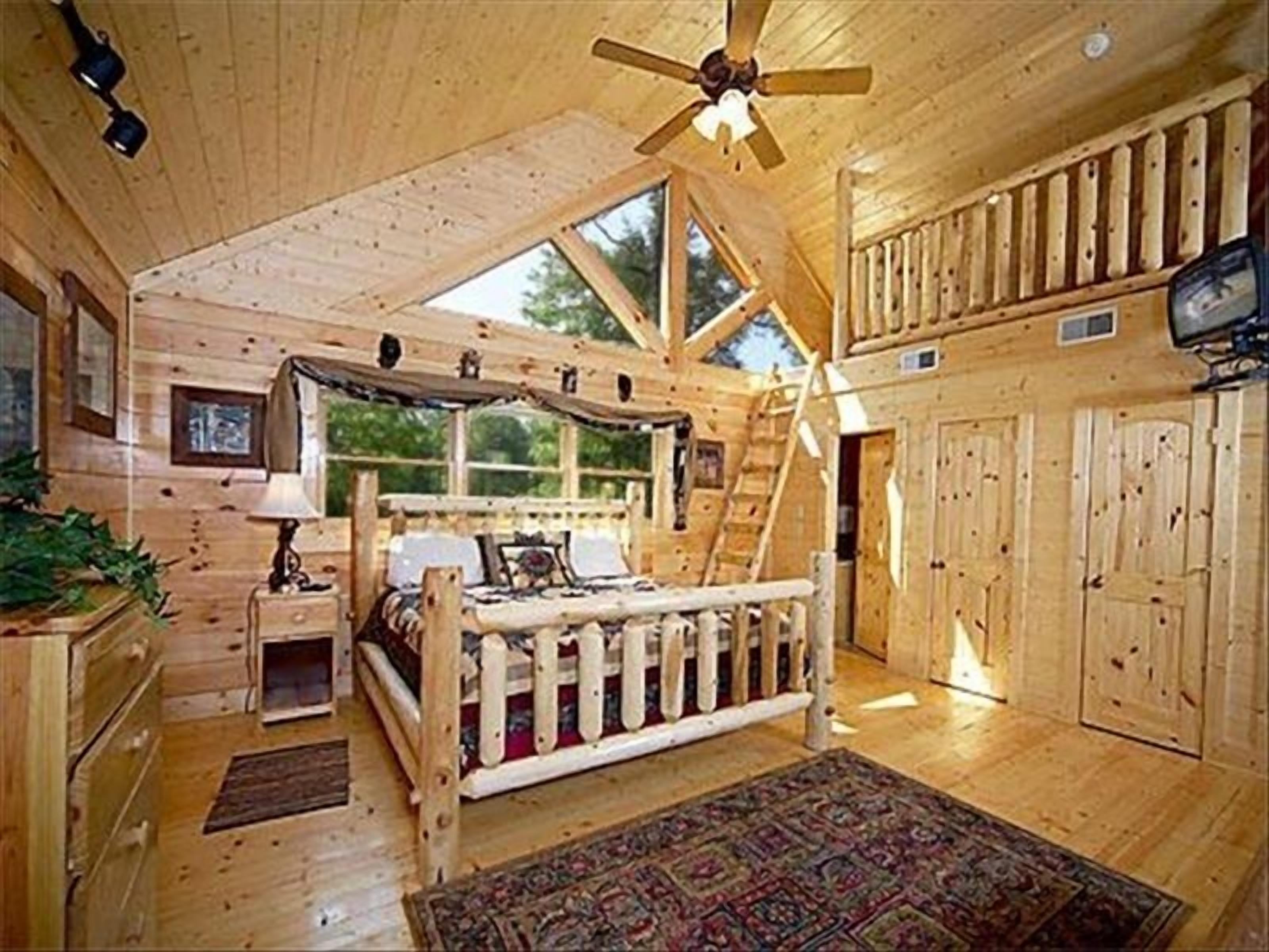 A log cabin interior with a loft bedroom, a wooden ceiling fan, and a large window.