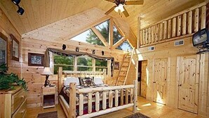 Een chaletinterieur met een slaapkamer op de zolder, een houten plafondventilator en een groot raam.