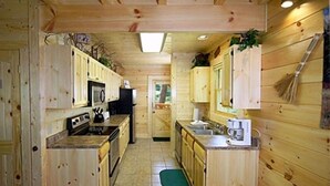 Een kleine keuken met houten kastjes, een magnetron, fornuis, spoelbak en een bezem die aan de muur hangt.