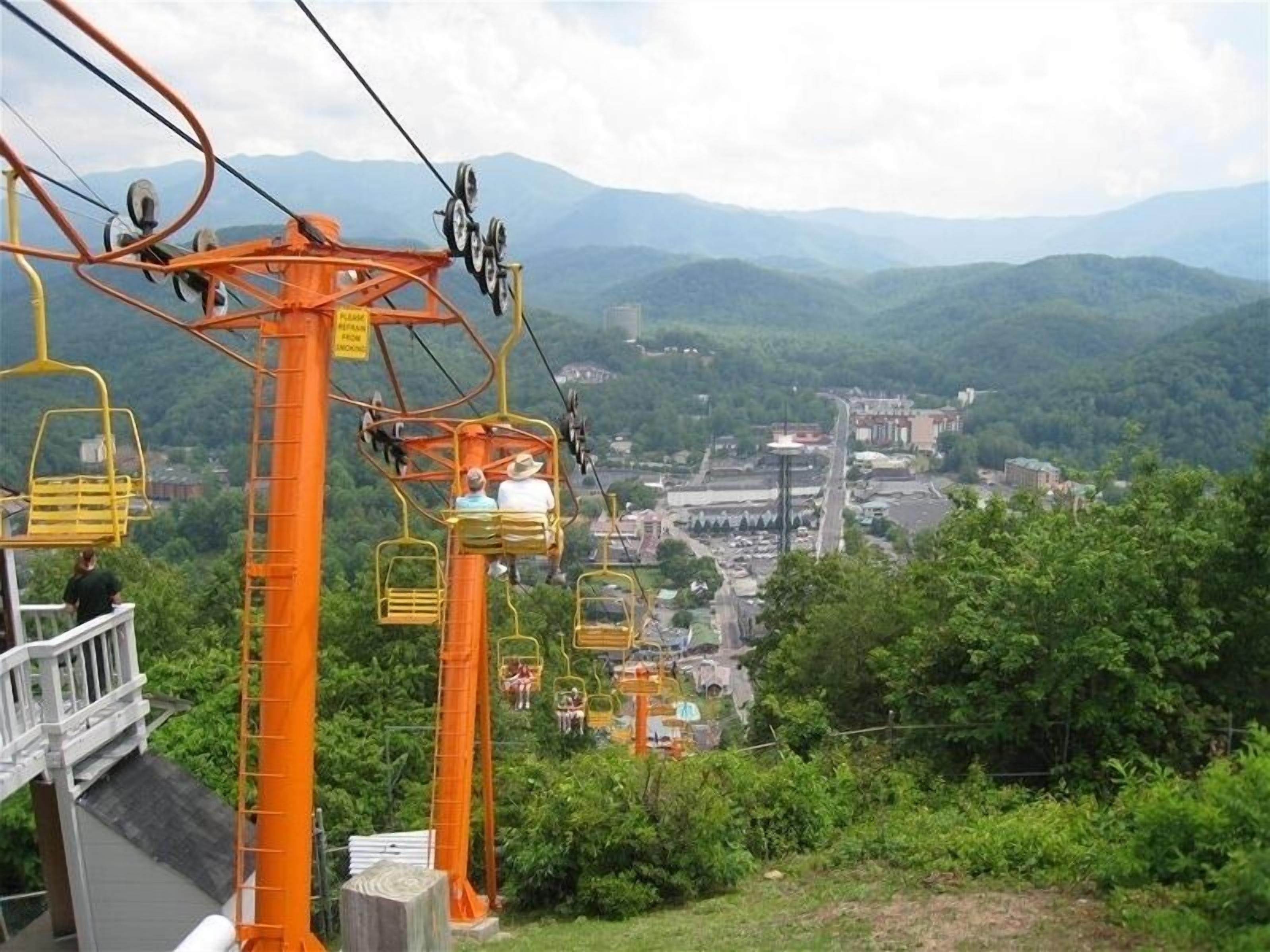 A cable car system with orange supports and yellow chairs, overlooking a valley with buildings and greenery.