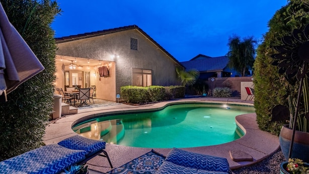 Una piscina con sillones de descanso, un patio con luces colgantes y una casa con garaje y ventanas visibles.