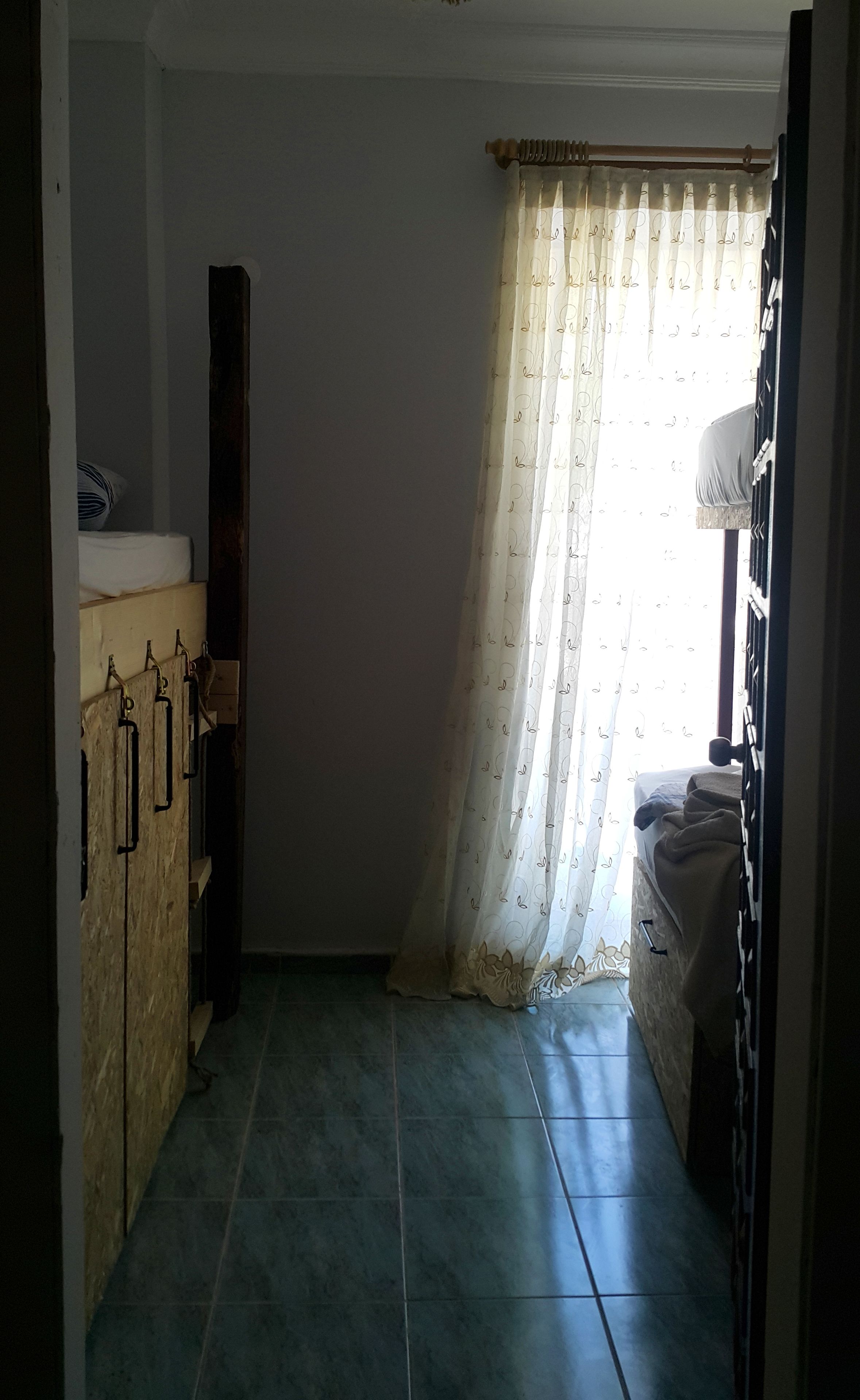 A room with a tiled floor, a wooden cabinet, a bed with a white blanket, and a window with a patterned curtain.