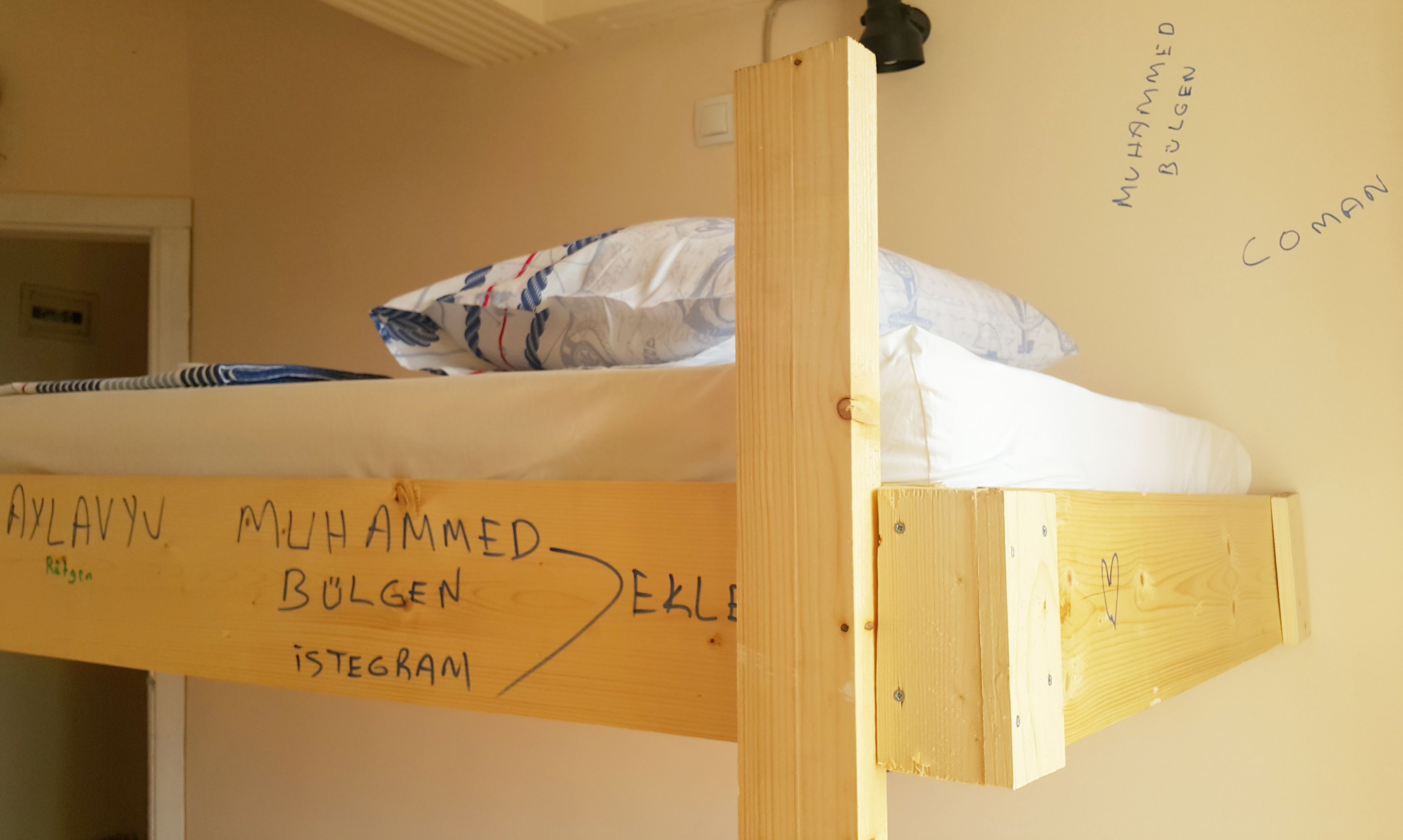 Un lit superposé avec une tête de lit en bois et un matelas recouvert d’une housse à motifs. Sur la tête de lit, on peut voir une pancarte manuscrite avec des noms et un identifiant de réseau social.