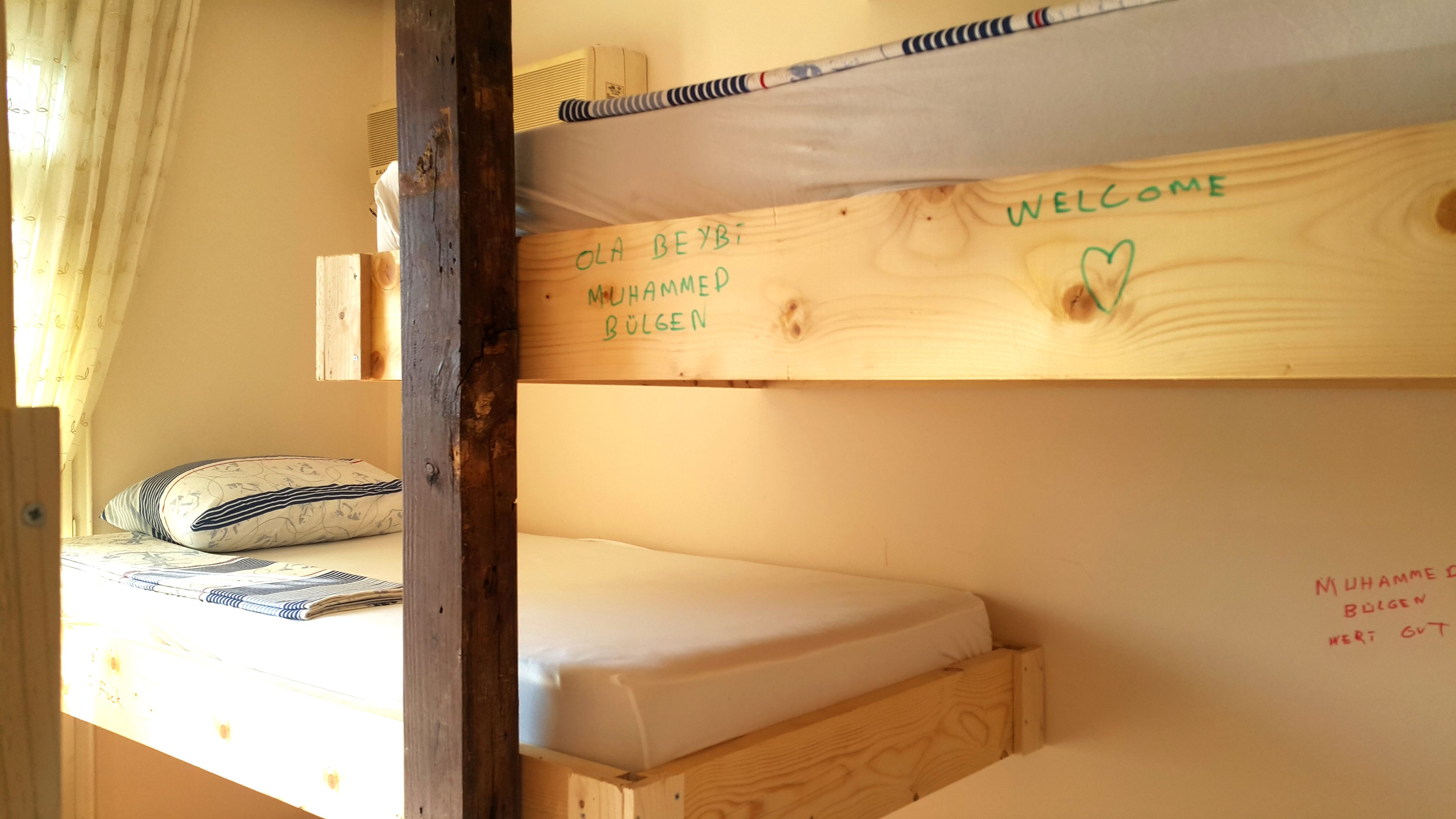 A bunk bed with a mattress and a pillow. There is a wooden beam with a 'WELCOME' sign and a name written on it.