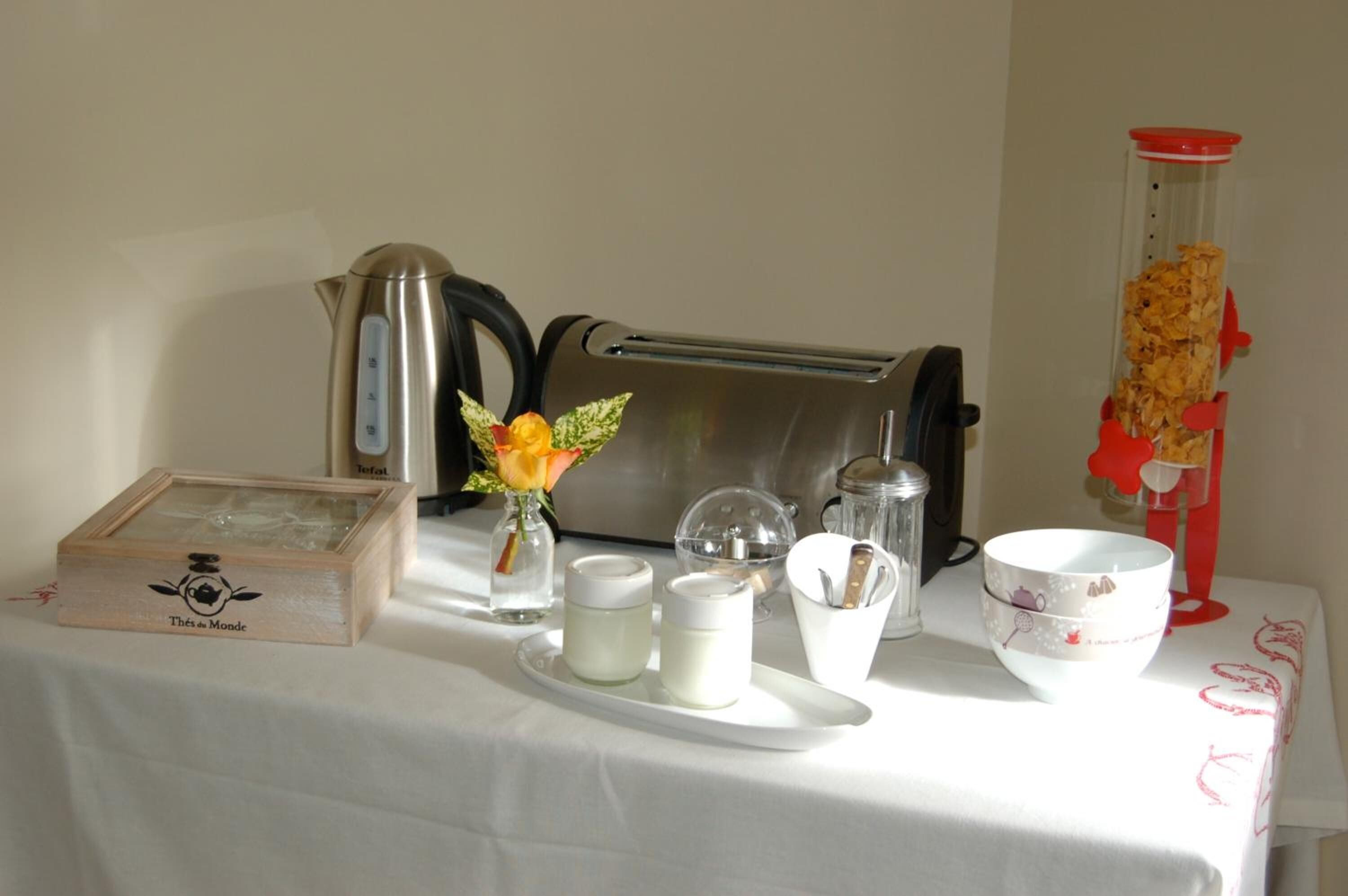 A table with a teapot, a toaster, a glass container with dried flowers, a small vase with a flower, two jars, a salt shaker, and a bowl.