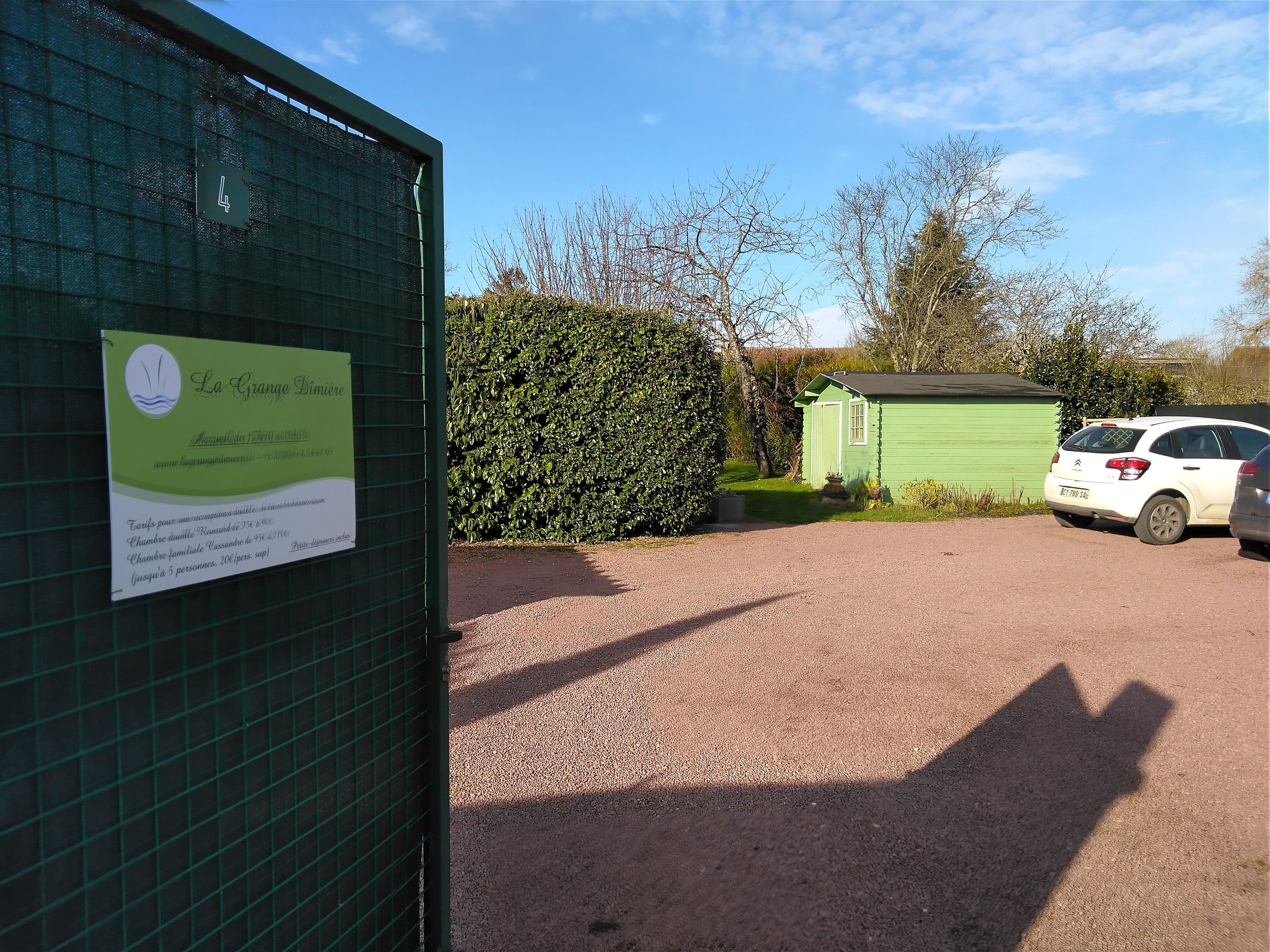 Une porte verte, sur laquelle est apposée une enseigne indiquant une maison d’hôtes nommée « La Grange Dinette », avec l’adresse de son site web et un numéro de téléphone. Un chemin de gravier mène à un bâtiment vert, près duquel est garée une voiture blanche.