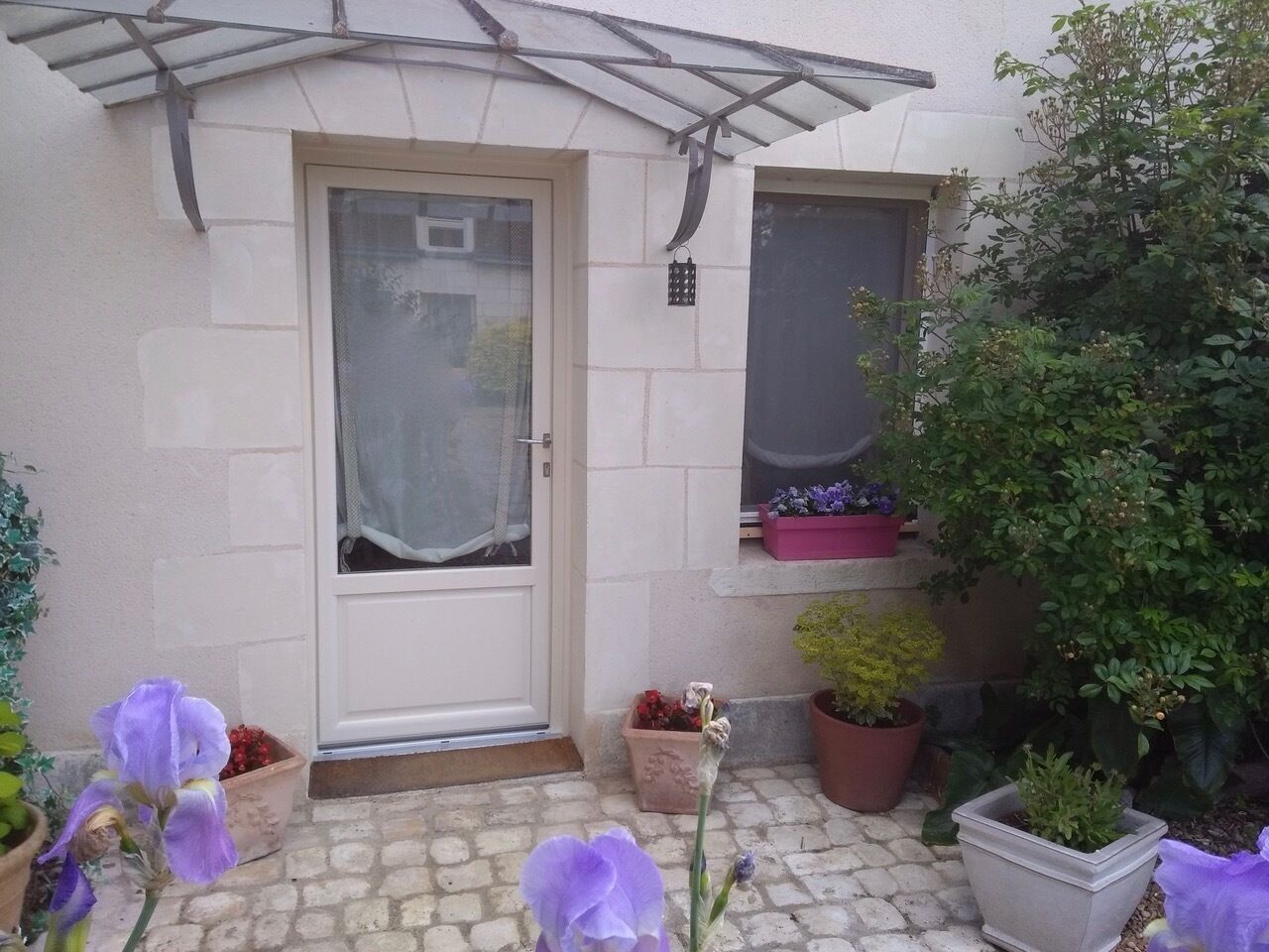 A small house with a front door, a window, and a garden with potted plants and flowers.