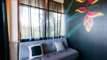 A room with a grey sofa, two pillows, a window with sheer curtains, and a wall mural of a tropical flower.