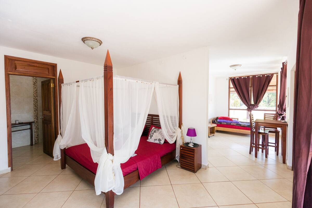 A four-poster bed with a red bedspread, a bedside table with a lamp, and a small wooden table with chairs in a bright room with tiled floors and a view of the outdoors.