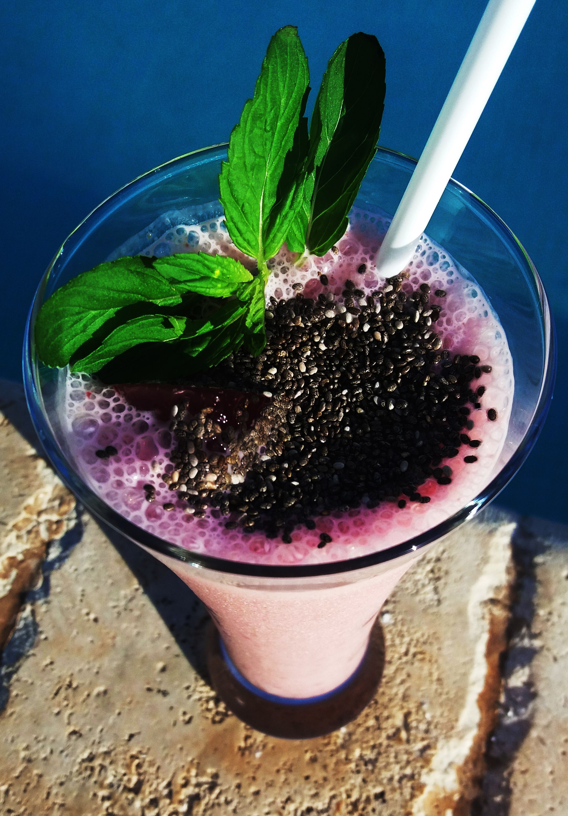 Un vaso de una bebida morada con semillas negras y hojas de menta, con una pajita blanca, sobre una superficie texturizada.
