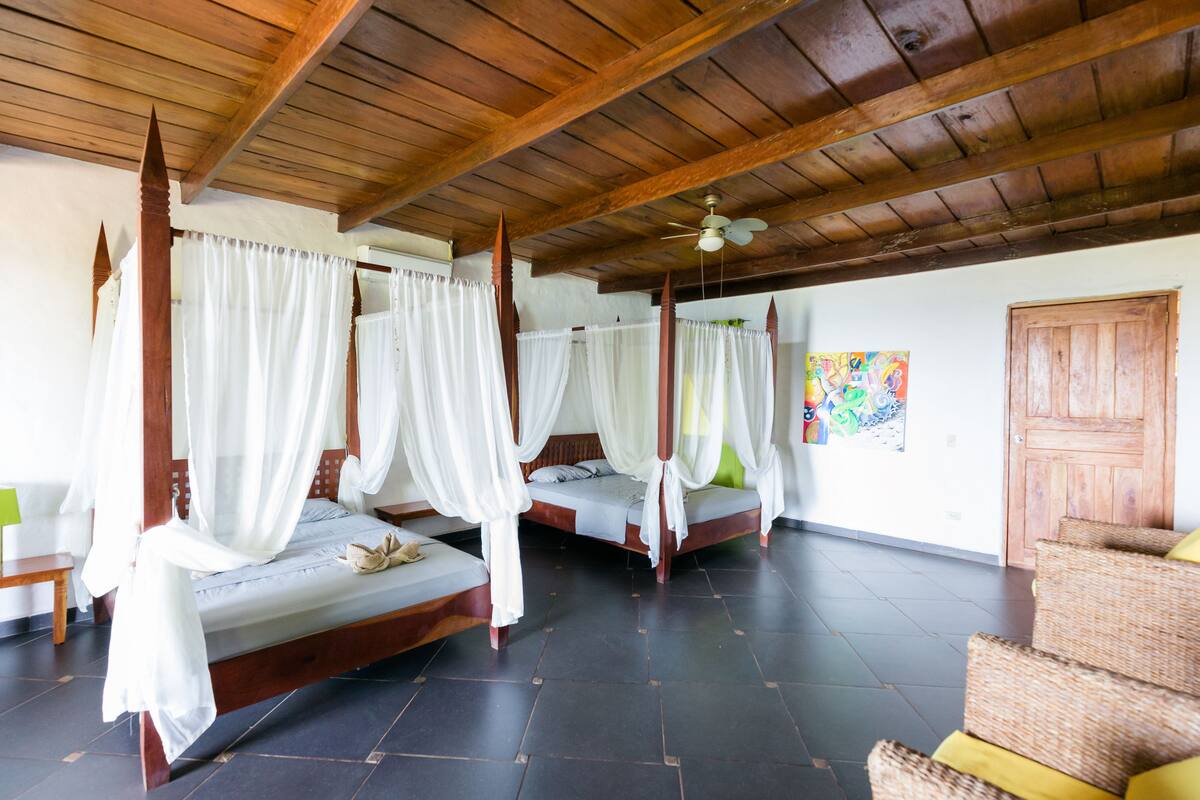 A room with four wooden four-poster beds, each with white curtains. There is a wooden door and a colorful painting on the wall.